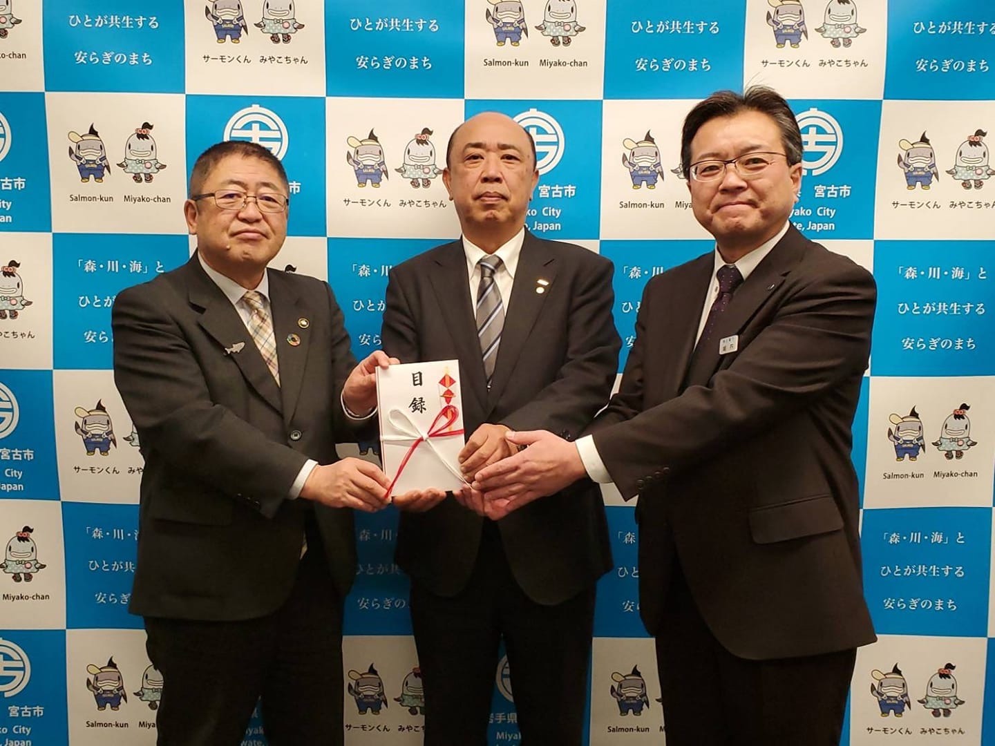 バックパネルの前で目録を一緒に持つ市長と男性2名が記念撮影をしている写真