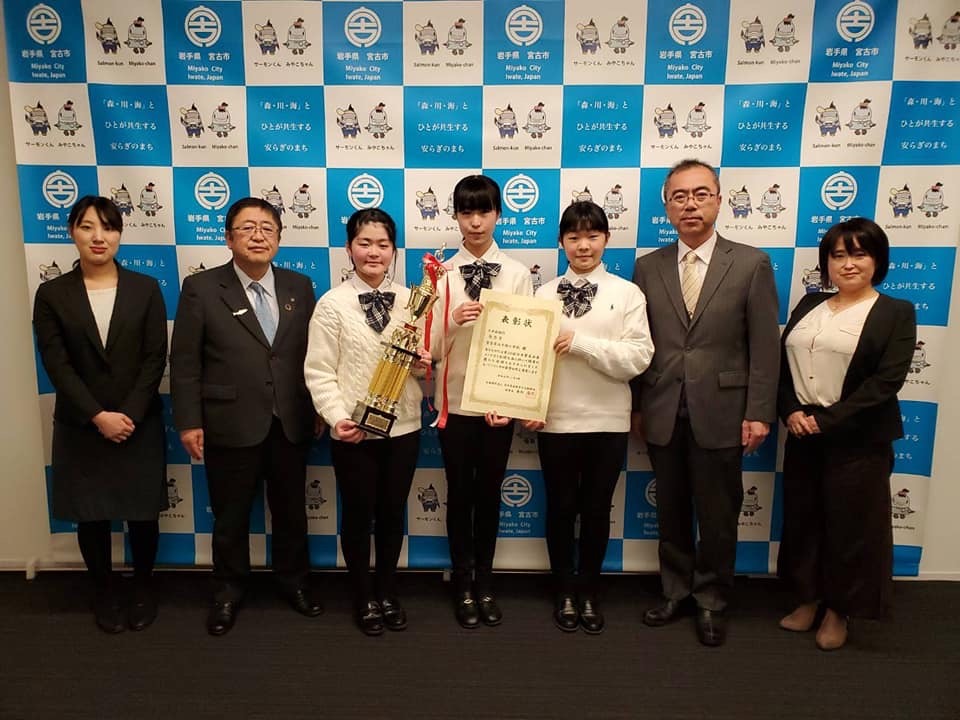 バックパネルの前で賞状とトロフィーを持つ制服を着た女子小学生3人を中心に市長を含む関係者4名が横一列に並ぶ記念写真