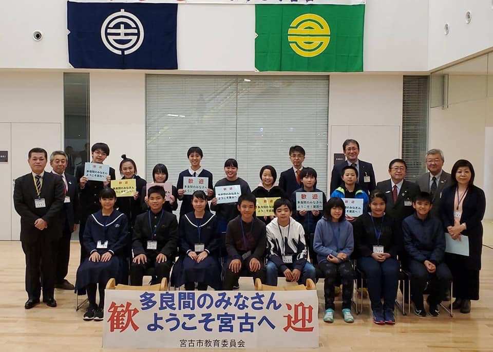 宮古市と多良間村の市町村旗が掲げられた会場で、「多良間のみなさんようこそ宮古へ」と書いてある看板の後方で1列目は椅子に座り2列目は立って両手に持ったプレートを胸の前に掲げている学生や関係者たちが並んでいる記念写真