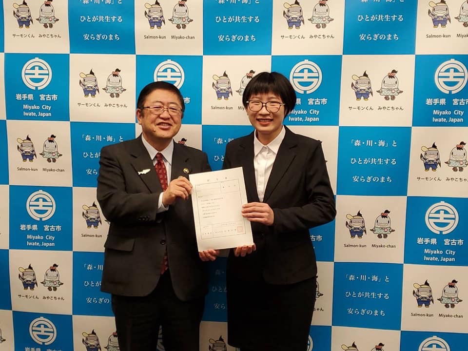 バックパネルの前で、辞令を一緒に持つ市長と女性が記念撮影をしている写真