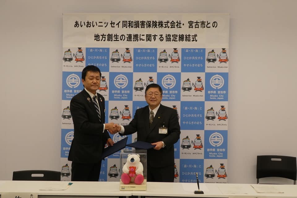 「あいおいニッセイ同和損害保険株式会社・宮古市との地方創生の連携に関する協定締結式」と書かれたバックパネルの前で、岩手支店長の佐藤氏と市長がそれぞれ協定書を持ち右手で握手を交わしている写真