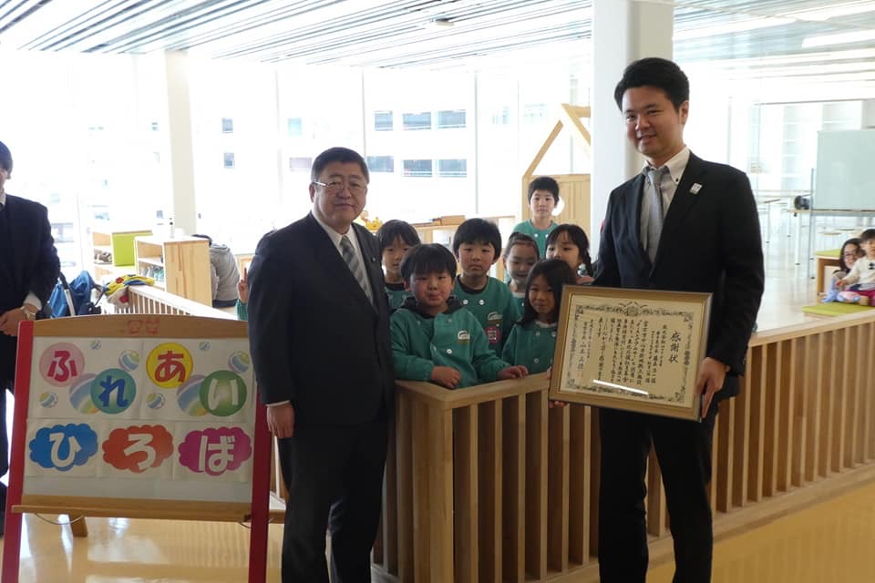 大きな窓ガラスが設置された「ふれあいひろば」と書かれた室内で、額入りの感謝状を受け取った東北支社岩手支店長の藤森氏と市長の後方に7名の子供たちが集まっている写真