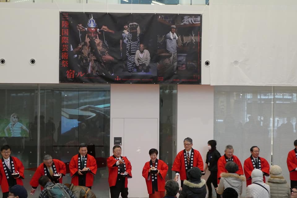 「三陸国際芸術祭 宿ル」と書かれたテーマと、宮古、八戸、大船渡の3枚のポスターを合体させた大きな幕が設置された会場で、赤色の法被を着た市長や関係者の方々が横一列に並んでいるオープニングセレモニーの様子の写真