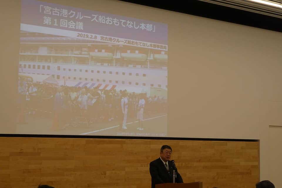 後方の壁に「「宮古港クルーズ船おもてなし本部」第1回会議」と書かれた会議資料が大きく映し出され、壇上に立つ市長が挨拶を行っている様子の写真