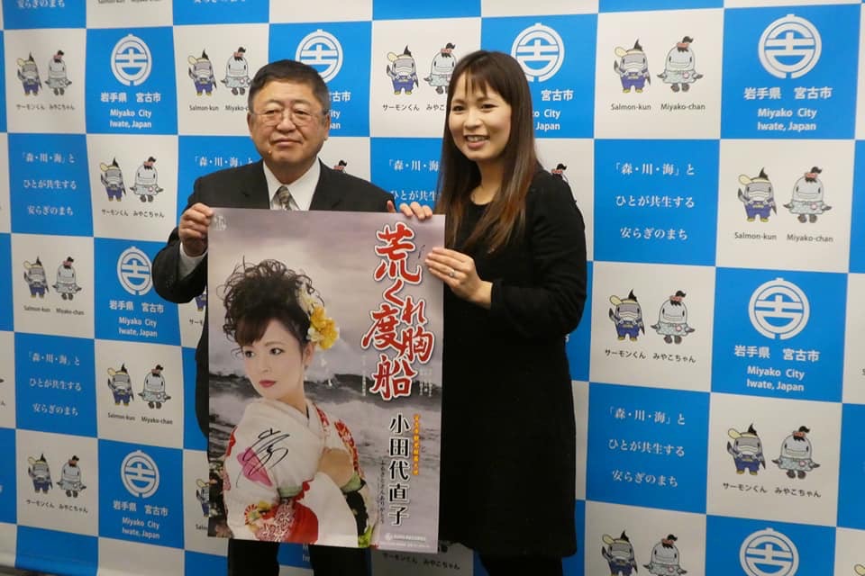 宮古市のバックパネルの前で、小田代直子さんと市長が新曲のポスターを一緒に持っている写真