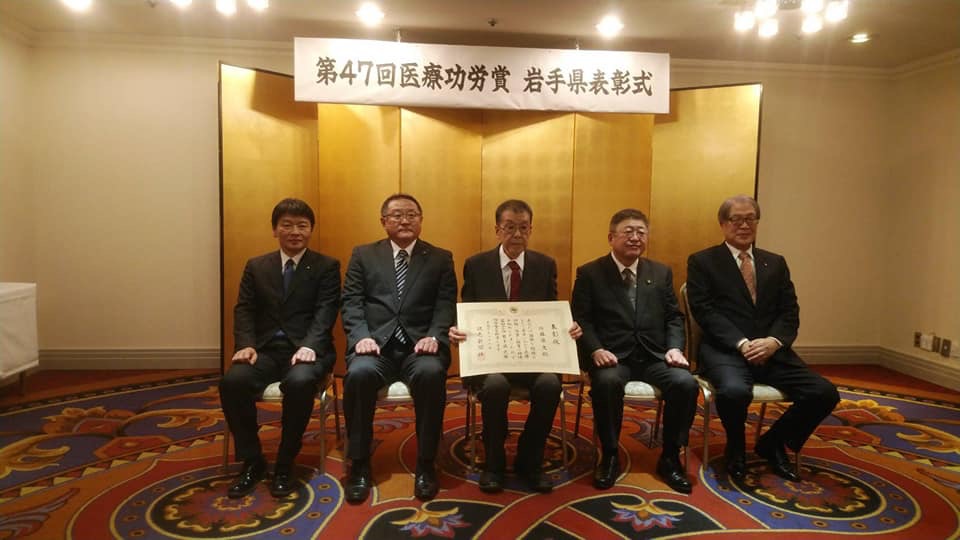「第47回医療功労賞 岩手県表彰式」と書かれた横幕と金屛風が設置された会場で、中央に賞状を持った後藤先生が座り、市長や3名の関係者の男性が横一列に並んでいる写真