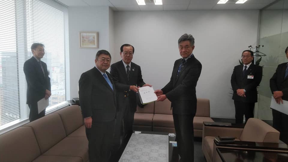 左側に大きな窓ガラスが設置されたKDDI株式会社東北総支社の室内で、テーブルを挟んで部長と向かい合って立つ市長と並んで立っている本田遠野市長が一緒に要望書を持ち、隅に3名の関係者が立っている写真