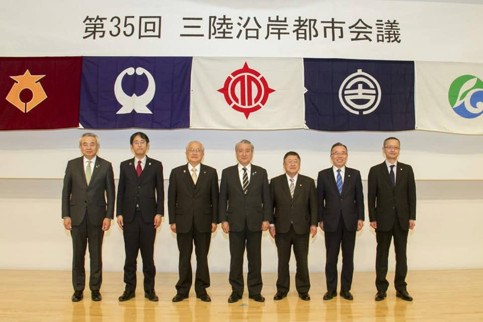「第35回三陸沿岸都市会議」と書かれた横断幕と各都市の旗が掲げられた会場に、市長を含め7名の首長が横一列に並んでいる写真