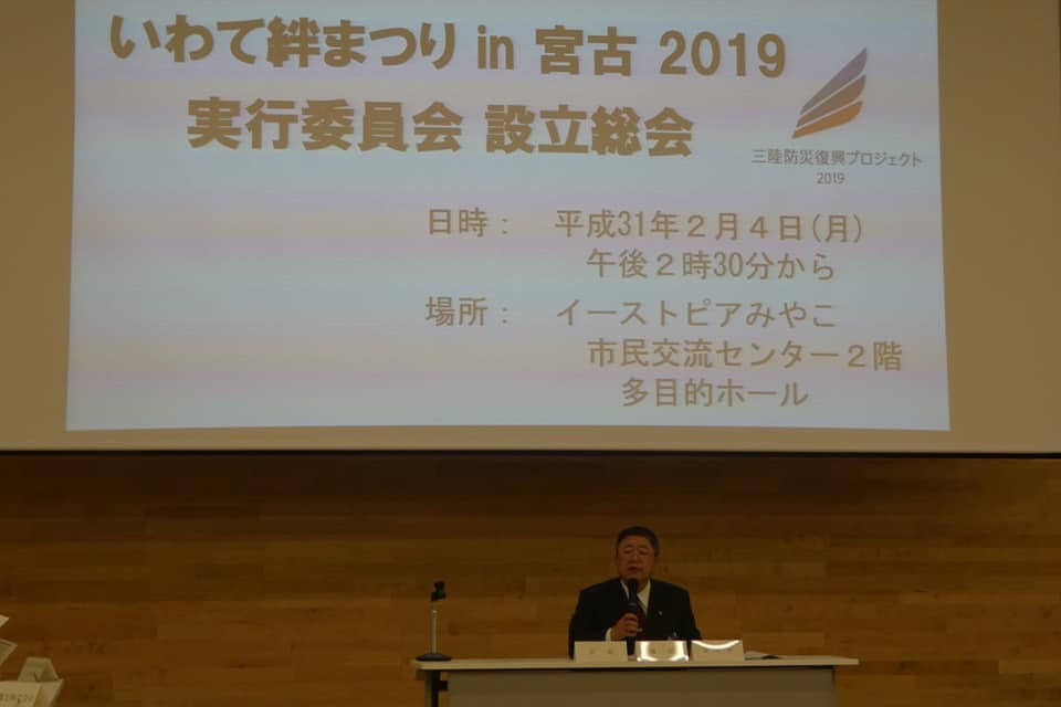後方のスクリーンに大きく「いわて絆まつりin宮古2019実行委員会設立総会」と書かれた資料の表紙が映し出され、長机の席に座った市長が右手にマイクを持ち話をしている写真