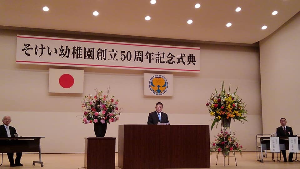 国旗と園旗に「そけい幼稚園創立50周年記念式典」と書かれた横断幕が掲げられたステージ左右に来賓者が座り、両サイドに綺麗に生けられた花が設置された壇上で挨拶を行っている市長の写真