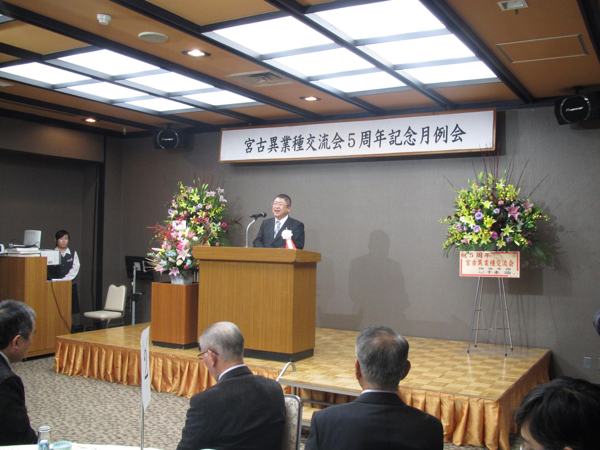 「宮古異業種交流会5周年記念月例会」と書かれた横断幕と花輪が置かれた舞台上で、演台に置かれたスタンドマイクに向かい話をする市長の写真