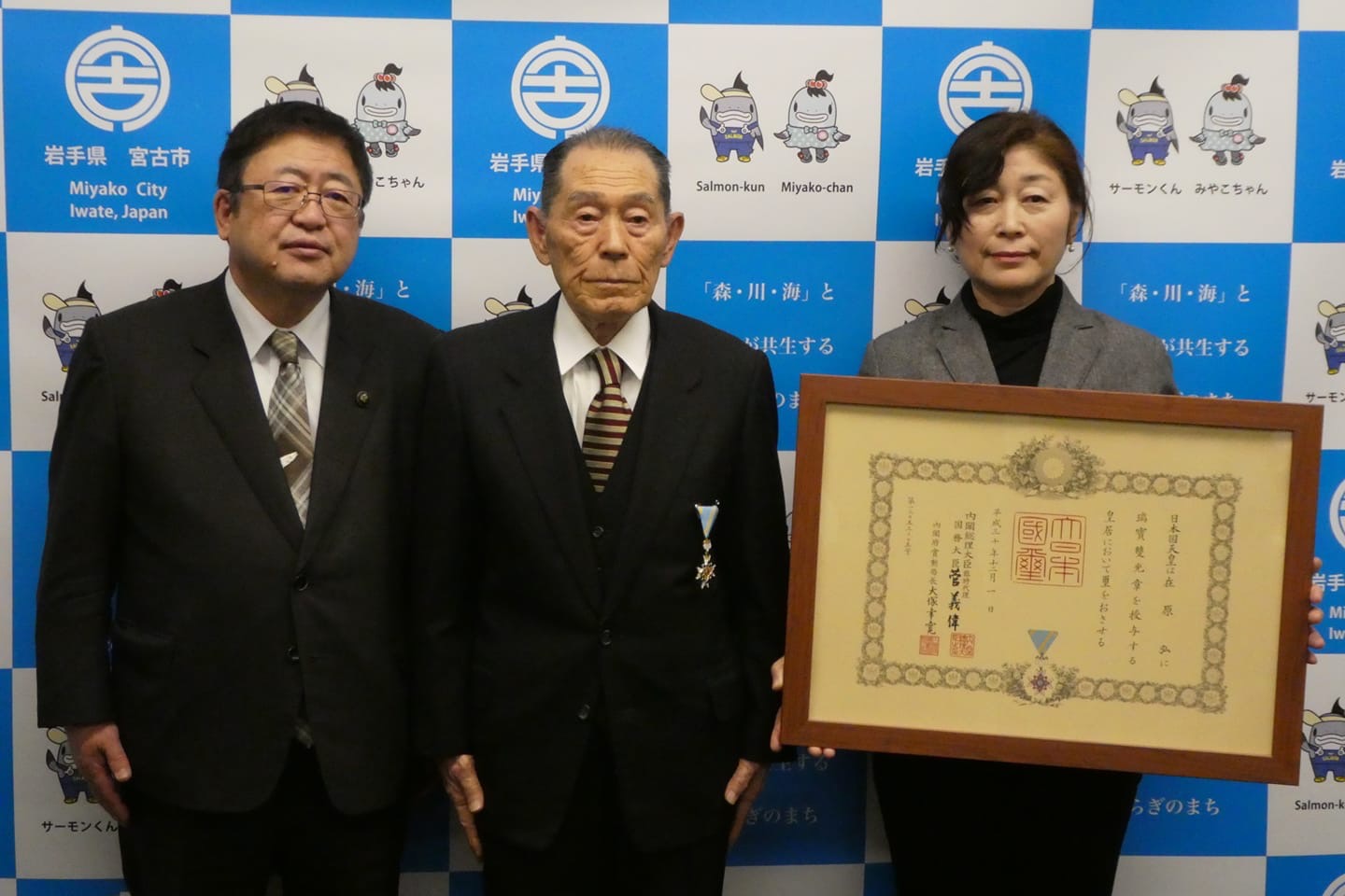 宮古市のバックパネルの前で、瑞宝双光章受章者の在原弘様を中央に右側に額入りの賞状を持った女性、左側に市長が横一列に並んでいる写真