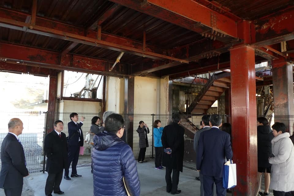 津波の被害にあい鉄筋の骨組みだけとなった震災遺構の建物下で、担当者から説明を受けている関係者の方々の写真