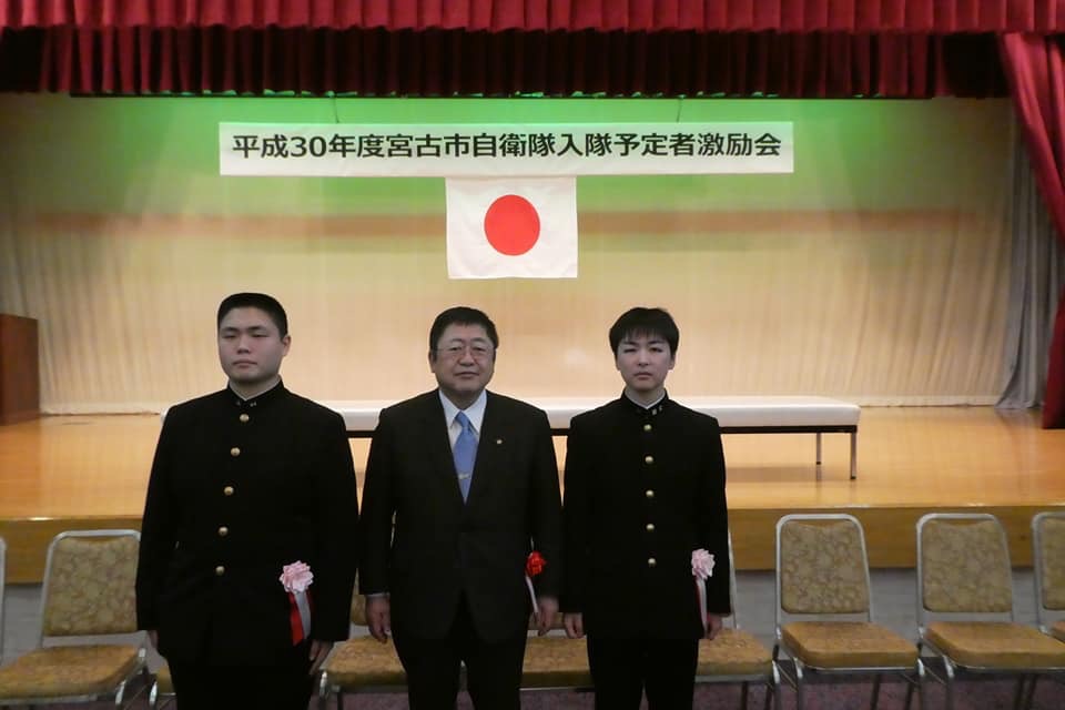 平成30年度宮古市自衛隊入隊予定者激励会」と書かれた横断幕と国旗が掲げられたステージ下で、市長を中央に学生服を着た柿本さんと白間さんと横一列に並んでいる写真