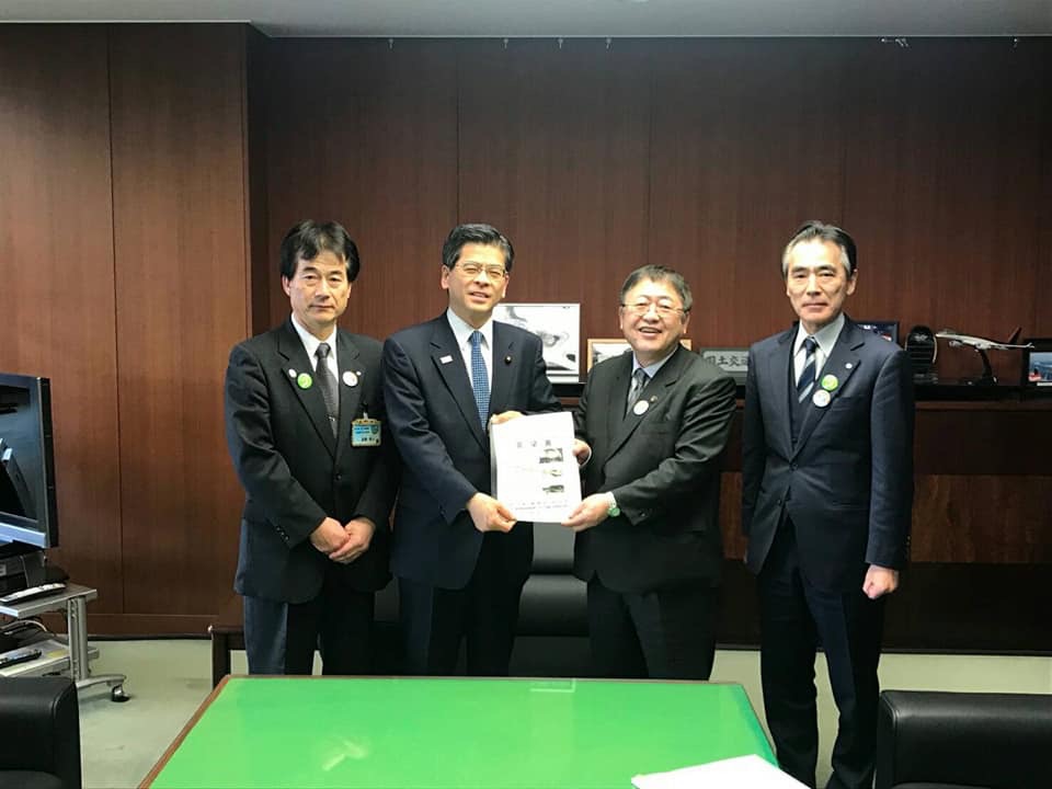 表面が黄緑色のテーブルや黒色の革張りの椅子が設置された室内で男性と市長が一緒に要望書を持ち、2名の関係者と横一列に並んでいる写真