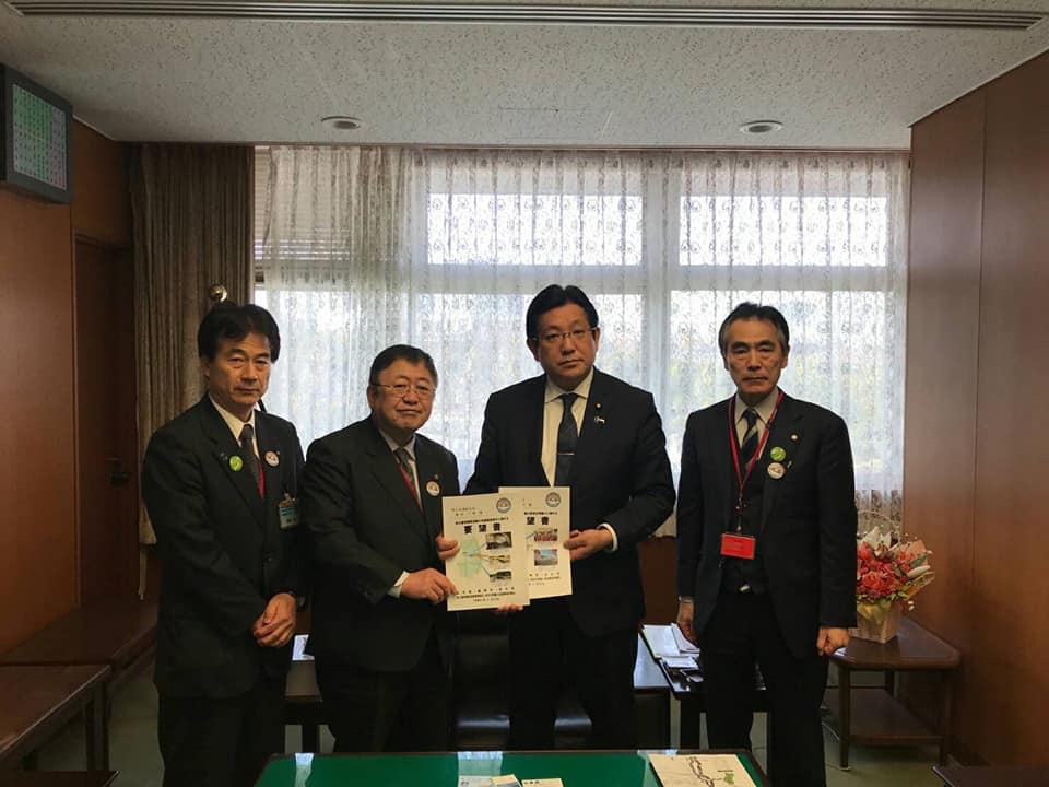 窓側に薄いレースのカーテンが敷かれた室内で、男性と市長が一緒に要望書を持ち、2名の関係者と横一列に並んでいる写真