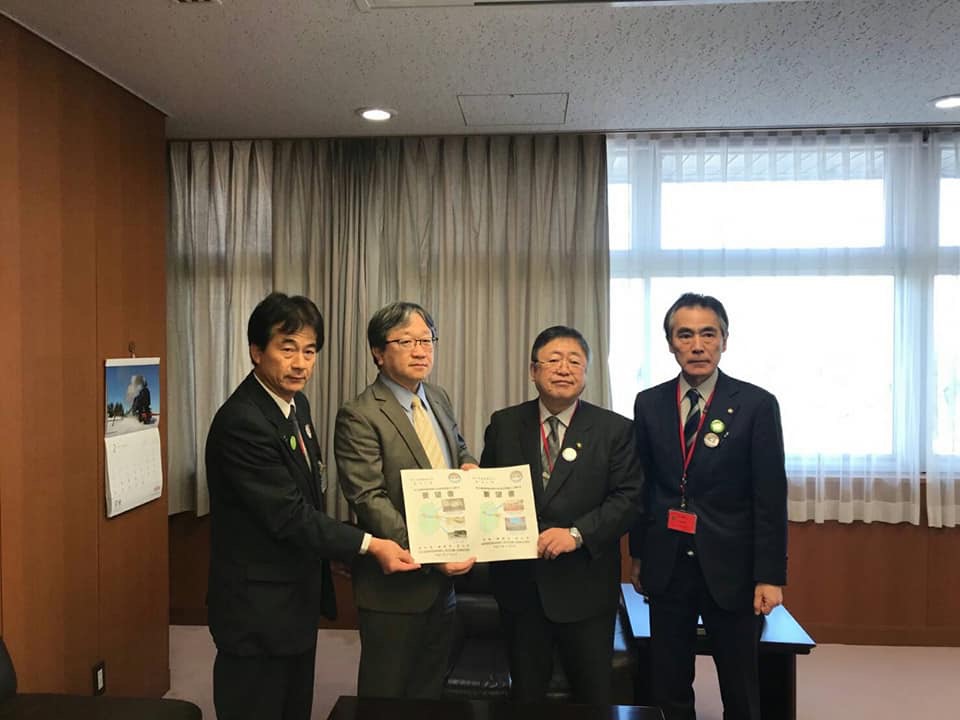 左側の壁にカレンダーが掛けられ窓側の薄いグレー色のカーテンが半分だけ締められている室内で、男性と市長が一緒に要望書を持ち、2名の関係者と横一列に並んでいる写真