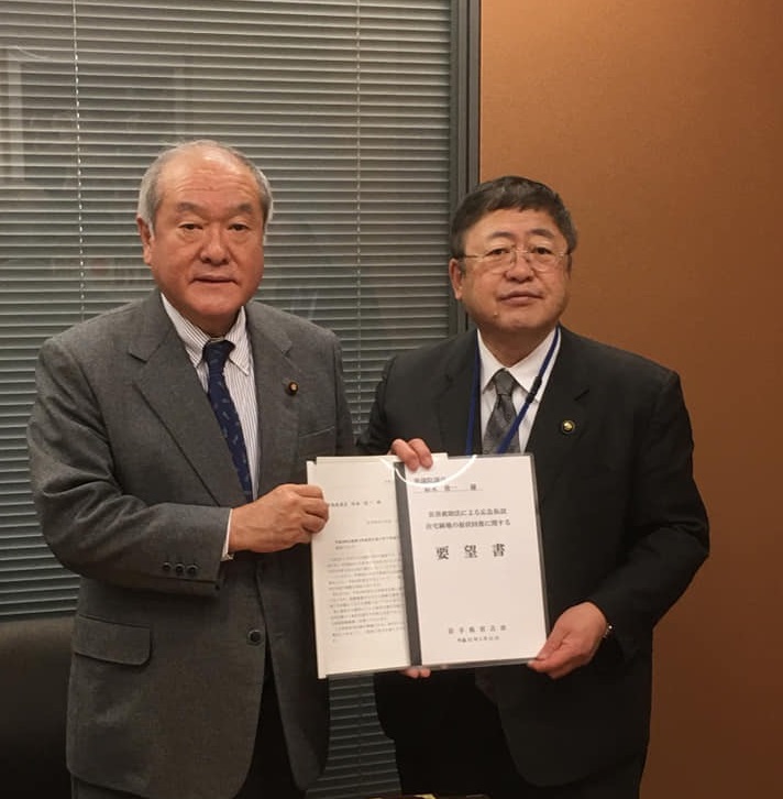窓にブラインドが設置された室内で、男性と市長が一緒に要望書を持っている様子をアップで撮影した写真
