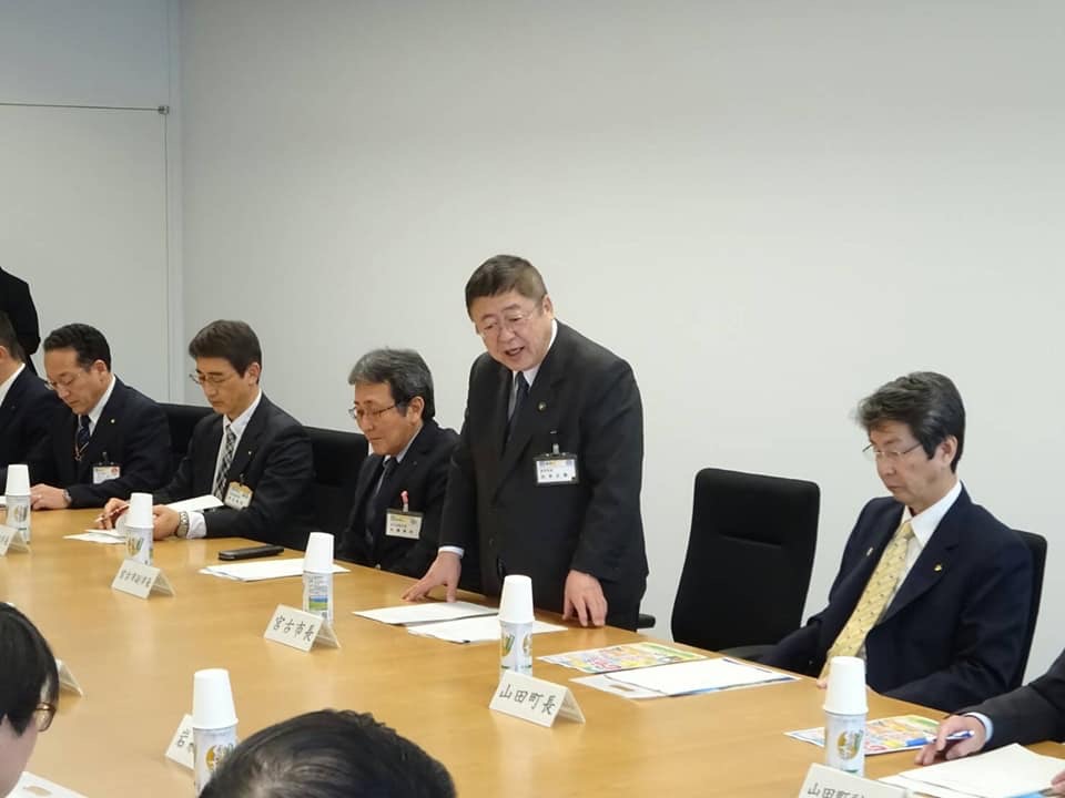 各市町村の代表が座っている会議室で、自席から立ち上がった市長が話をしている様子の写真