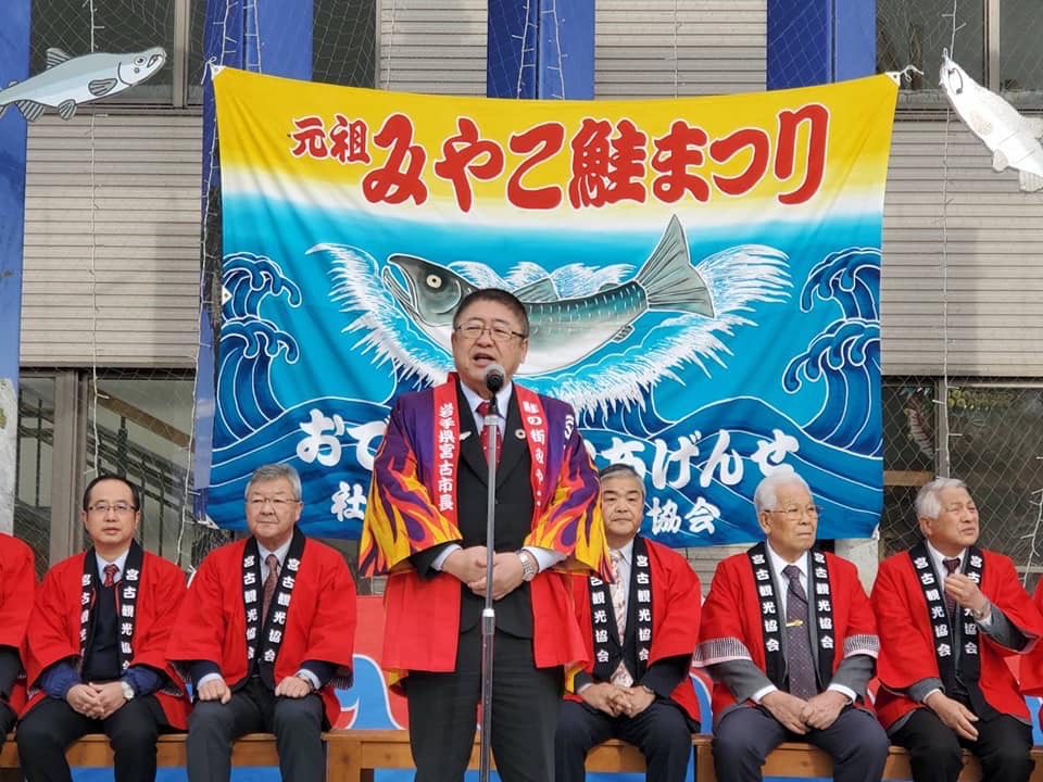 「元祖みやこ鮭まつり」という文字と絵が描かれた旗の前で法被を着て椅子に座る関係者たちと、スタンドマイクの前で両手をお腹の前で組み話をする様子の市長の写真