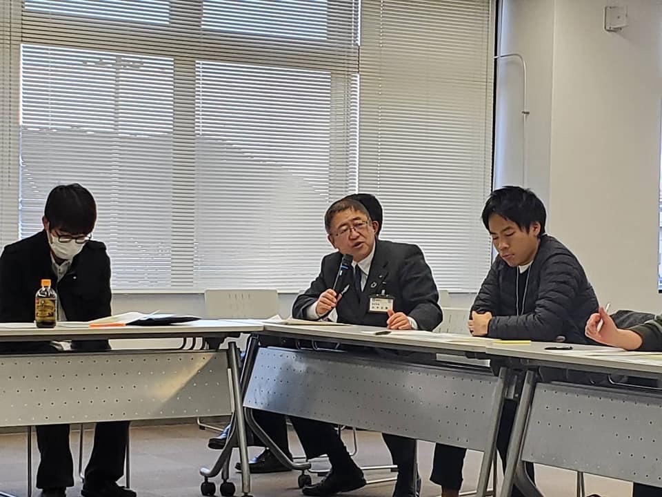 L字に並んだ席に着き、マイクを持って話をする市長とその周りで座って手元の資料を見ている様子の関係者たちの写真