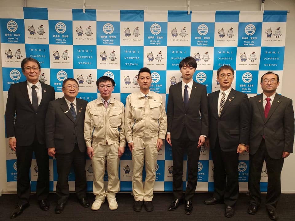 バックパネルの前で横一列に並ぶ市長を含めた5名のスーツを着た男性と2名の作業着をきた男性の記念写真