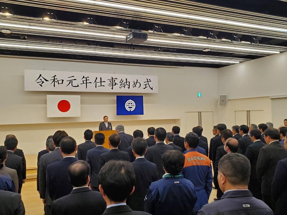 「令和元年仕事納め式」と書かれたパネルと国旗、宮古市の市町村旗が吊るされた部屋で、演台の前で話をする様子の市長と整列して話を聞く職員たちを写した写真