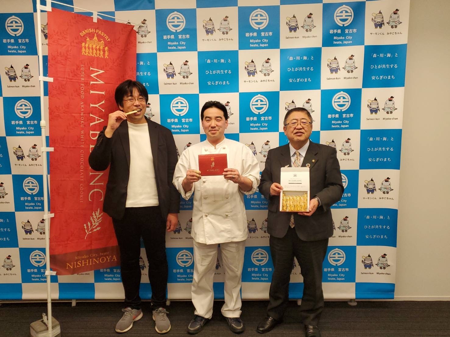 バックパネルの前で商品を持つ市長と男性2名を写した記念写真