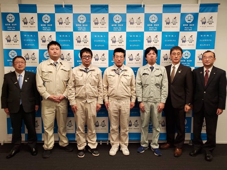 作業服を着た派遣職員4名と、市長、2名のスーツを着た関係者の男性が横一列に並ぶ記念写真