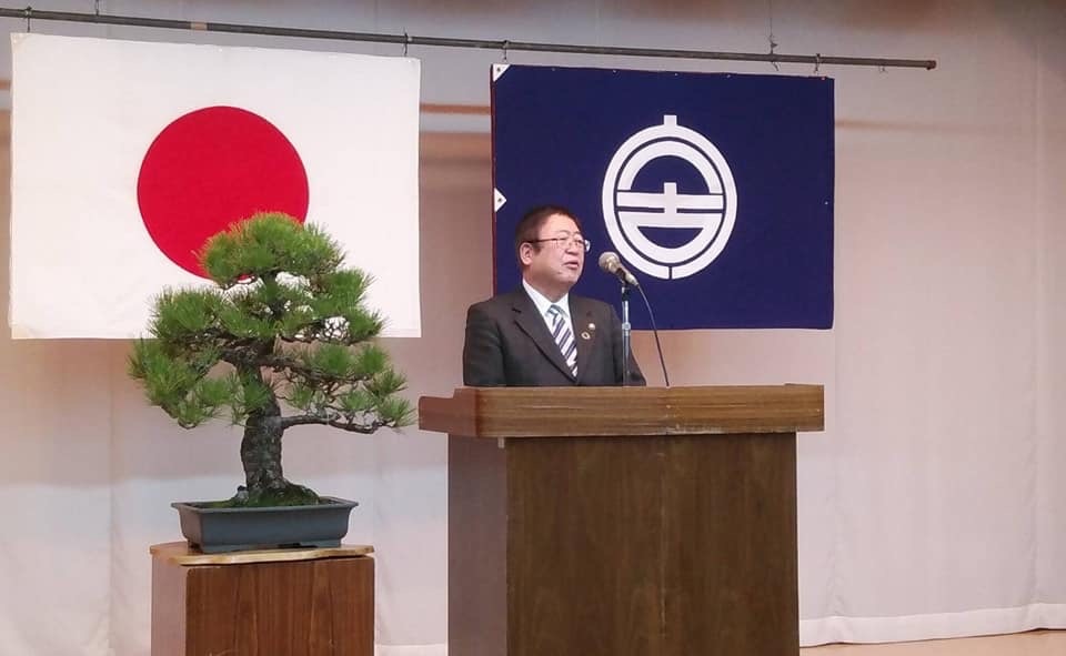 日本国旗と宮古市の市町村旗が吊るされ、台に置かれた大きな盆栽の横の演台で、マイクに向かって話をしている市長の写真