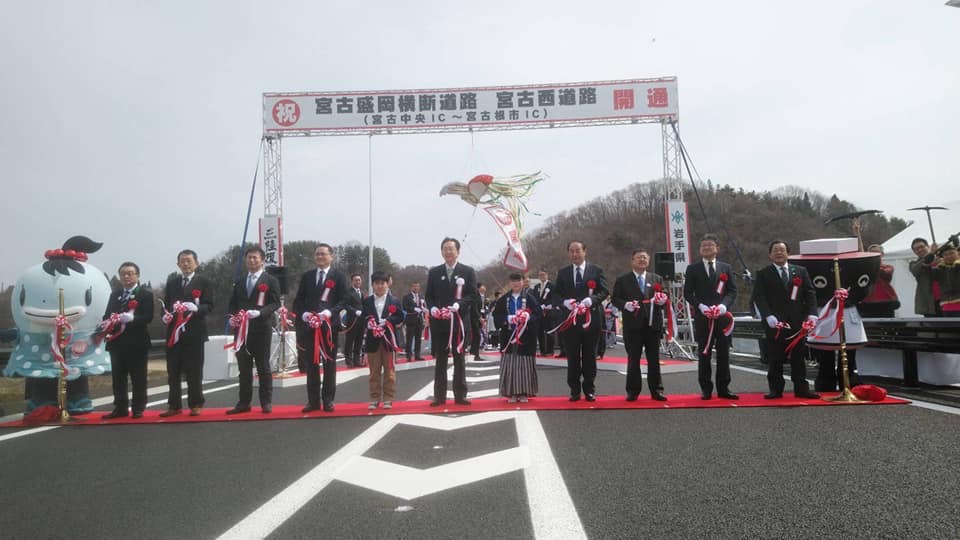 道路を横断するように敷かれたレッドカーペットの上でテープカットをする様子の市長や関係者の男性8名と男の子1名、女の子1名、みやこちゃんとわんこきょうだいの写真