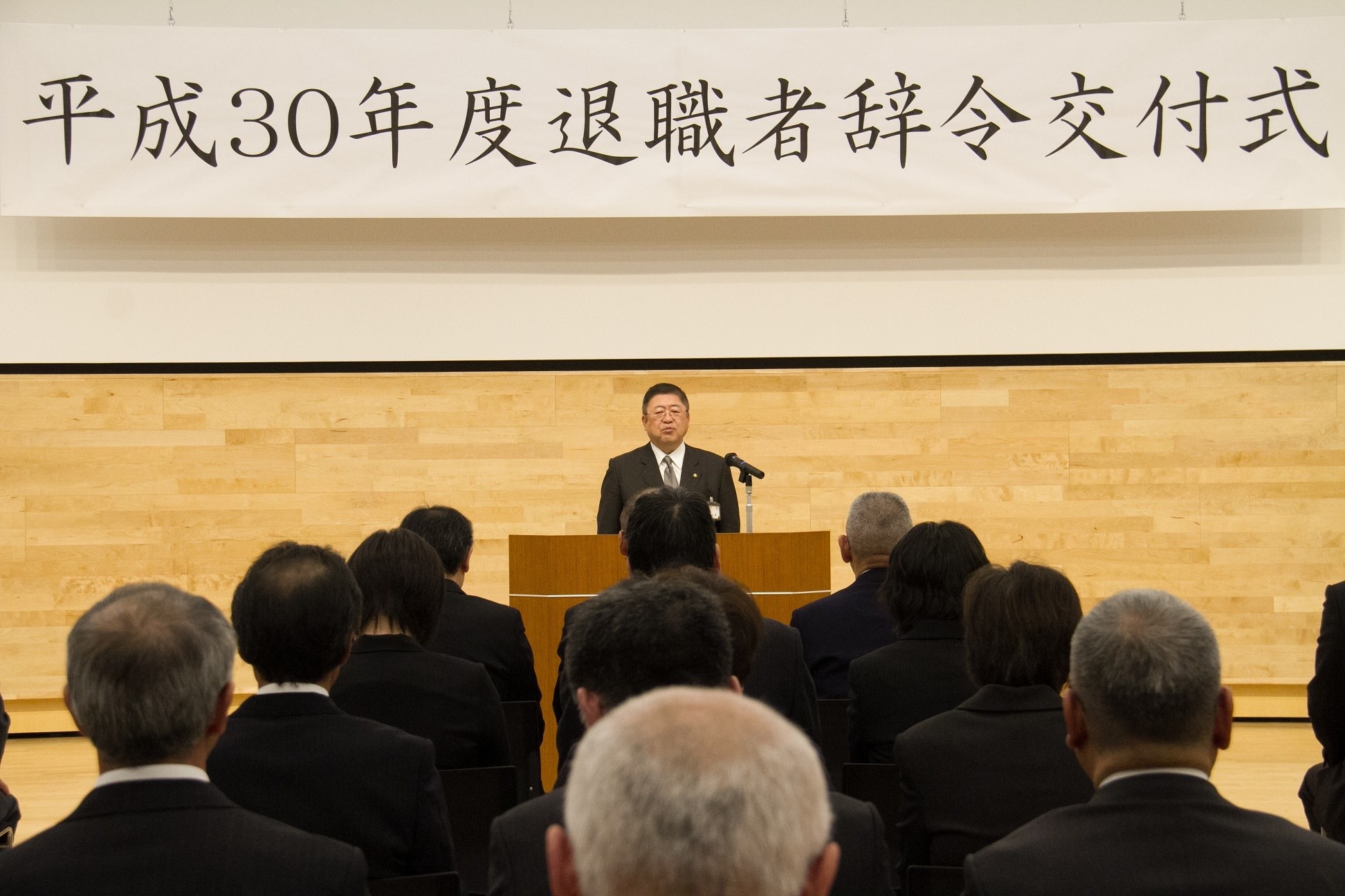 「平成30年度退職者辞令交付式」と書かれた横断幕が吊るされた会場で、整列する職員の前で演台に置かれたマイクに向かい話をする様子の市長の写真