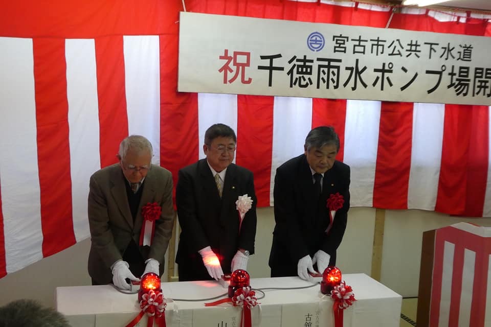 紅白幕の前にあるテーブルに置かれた赤いランプのスイッチを入れる様子の市長と関係者の男性2名の写真