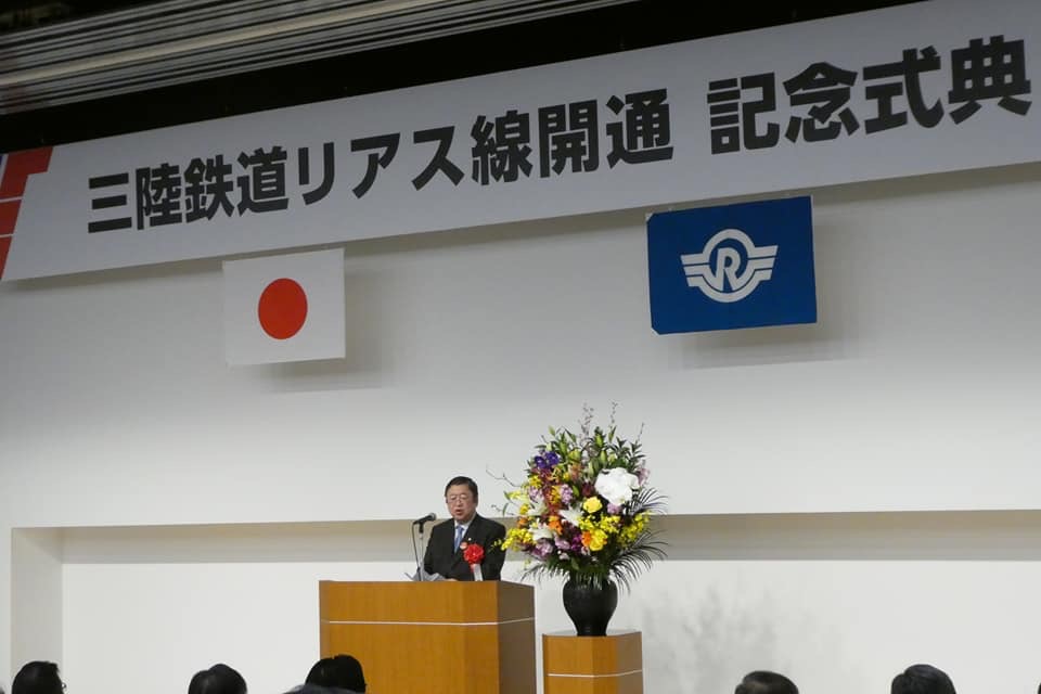 「三陸鉄道リアス線開通 記念式典」と書かれた横断幕、日本国旗、三陸鉄道の旗が掲げられた会場で、演台に置かれたマイクに向かい話をする様子の市長