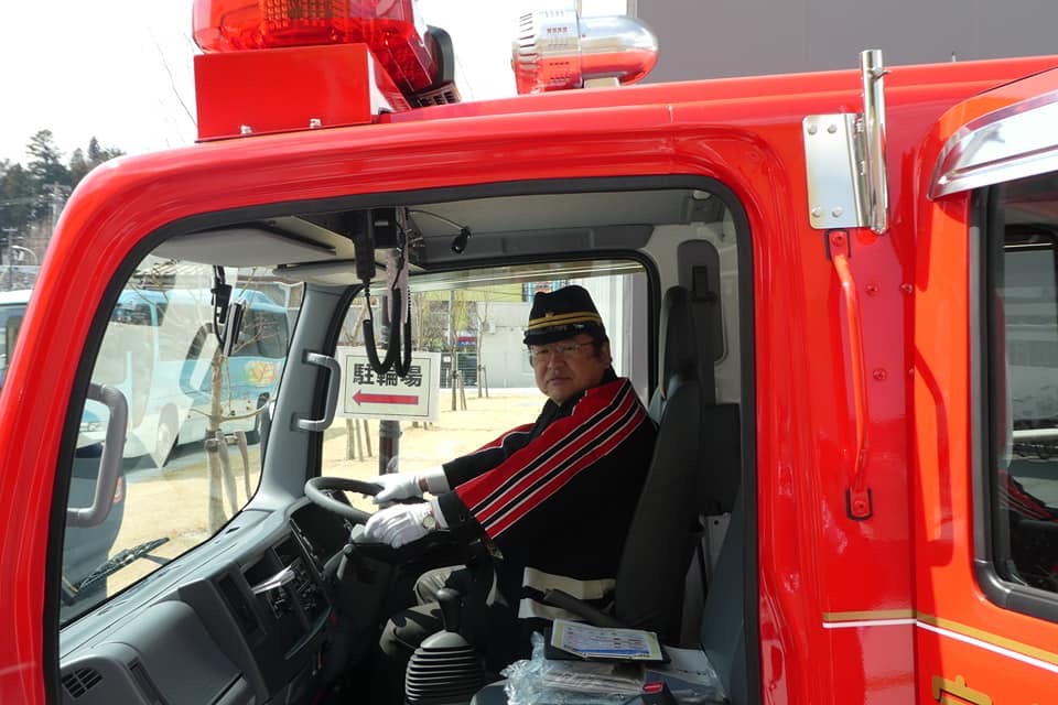 消防の法被と帽子を身に着け消防車に乗りハンドルを両手で握っている市長の写真