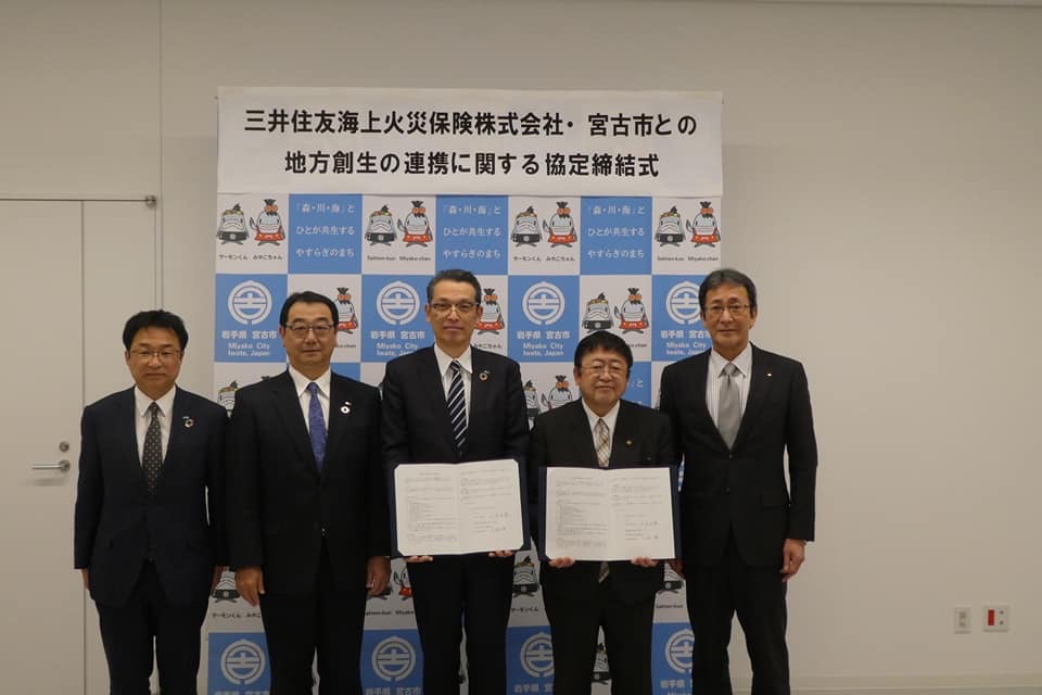 「三井住友海上火災保険株式会社・宮古市との地方創世の連携に関する協定締結式」と書かれた紙が貼られたバックパネルの前で、締結書を持つ市長と男性、その両側に3名の関係者の男性が並ぶ記念写真