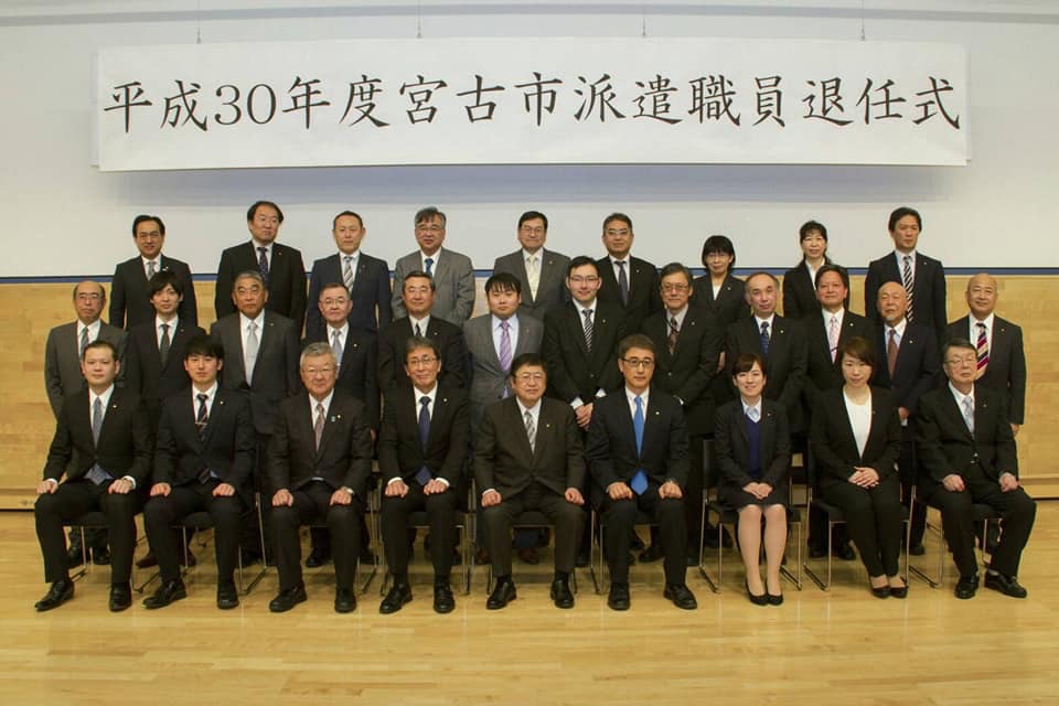 「平成30年度宮古市は県職員退任式」と書かれた横断幕が吊るされた会場で、1列目に椅子に座る市長と関係者や派遣職員の方々8名、2列目に12名、3列目に11名が並んでいる記念写真