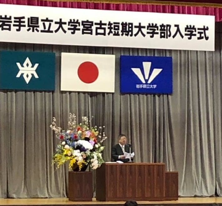「岩手県立大学宮古短期大学部入学式」と書かれた横断幕が掲げられた舞台上で、色とりどりの花が活けられた花瓶が置かれたテーブルの横にある演台に置かれたマイクに向かい話をする様子の市長の写真
