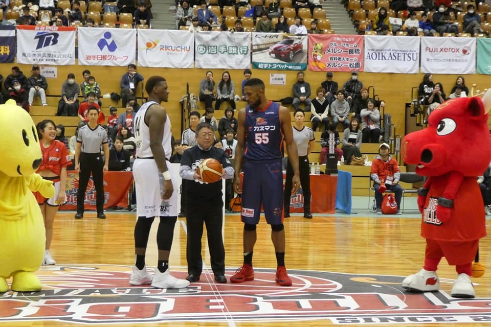 試合コートでユニフォームを着た2名の黒人選手の間にバスケットボールを持って立っている市長の写真
