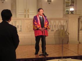 赤い法被を着た市長がお腹の前で両手を組みスタンドマイクに向かって話をしている様子の写真
