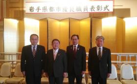 「岩手県都市優良職員表彰式」と書かれた横断幕が吊るされ、金屏風が設置された会場で市長と3名の男性が横並びになっている記念写真