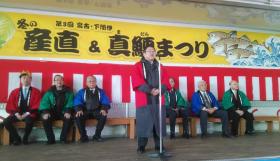 「第3回 宮古・下閉伊 冬の産直&真鱈まつり」と書かれた横断幕が吊るされ、紅白幕が貼られた壁際に置かれた椅子に法被を着た関係者の男性6名が座り、その前に置かれたスタンドマイクの前で話をする市長の写真