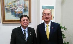 額縁に入った絵画が飾られた壁の前で並んで写る市長と男性の記念写真