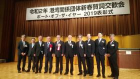 「令和2年 港湾関係団体新春賀詞交歓会 ポート・オブ・ザ・イヤー2019表彰式」と書かれた横断幕が吊るされた舞台上でスーツを着た男性10名が横一列に並び記念撮影をしている写真