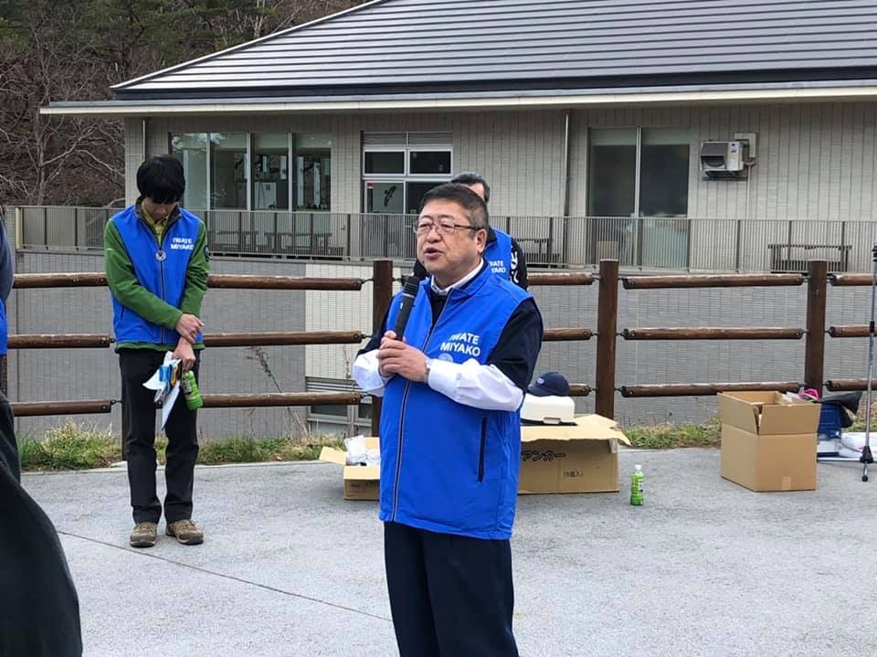 青色のベストを着て両手でマイクを持ち話をする様子の市長の写真