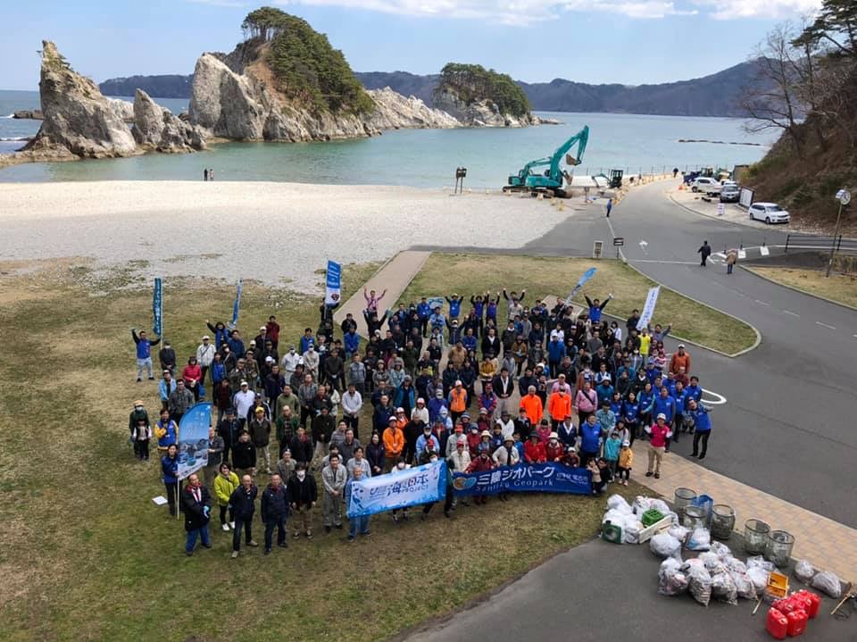 清掃された浄土ヶ浜と横断幕や旗を掲げ集合している大勢の参加者と回収されたごみの入った複数のごみ袋を上空から写した写真