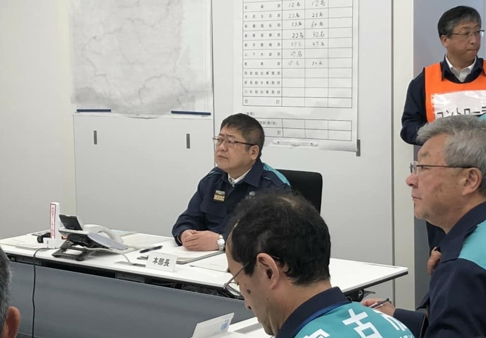 電話機が置かれたテーブルの席に着き、組んだ両手をテーブルの上に置き前方を見つめる様子の市長の写真