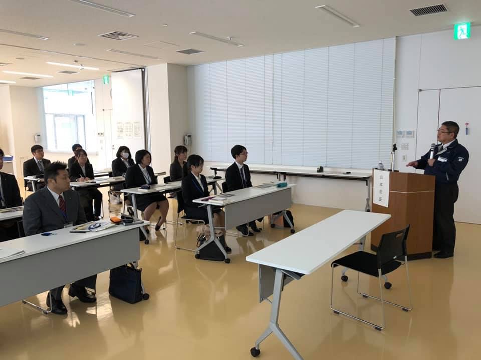 机がスクール形式に並べられ、1台に2名ずつスーツを着た宮古市新採用職員の方々が座り、演台の前でマイクを持ち話をしている市長を見ている様子の写真