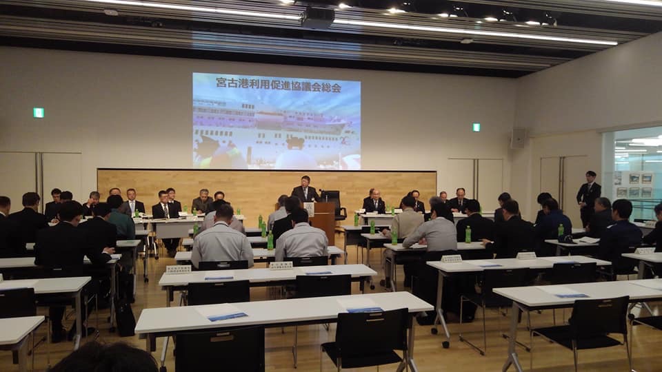 プロジェクターで画像が投影された壁の前方で演台に向かい立つ市長や、会場に並べられたテーブルの席に着く参加者たちの写真