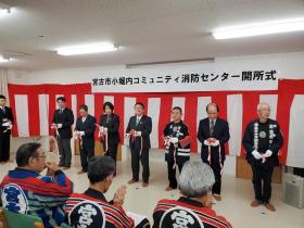 宮古市小堀内コミュニティ消防センター開所式と書かれた幕の下の壁に紅白の幕を張り、 それを背景にして、市長をはじめ、6人で消防団の法被を着た分団員の前でテープカットをした写真